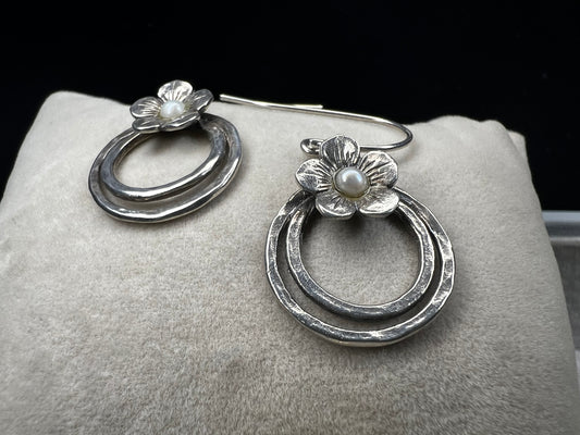 These sterling silver earrings showcase a flower design with a lustrous pearl at its center, elegantly positioned atop a double hoop. The combination offers a refined aesthetic, perfect for adding a sophisticated touch to any ensemble.