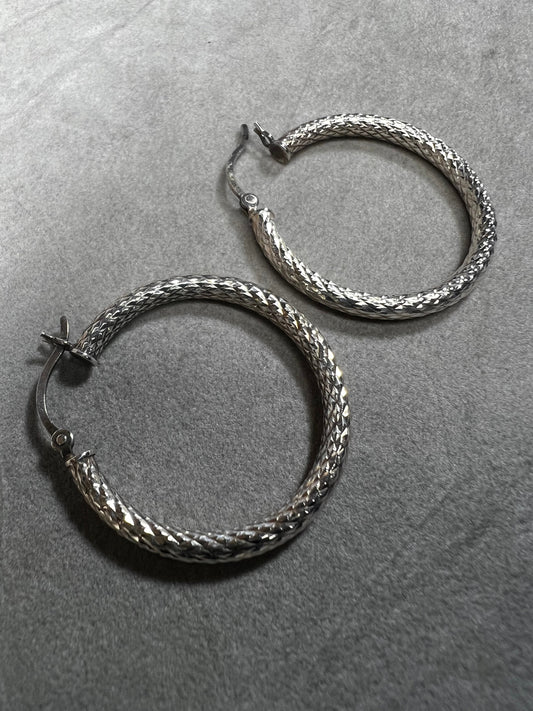 These sterling silver hoop earrings feature a diamond-cut texture that adds subtle shimmer and dynamic movement. Their moderate size balances statement presence with comfortable wearability.