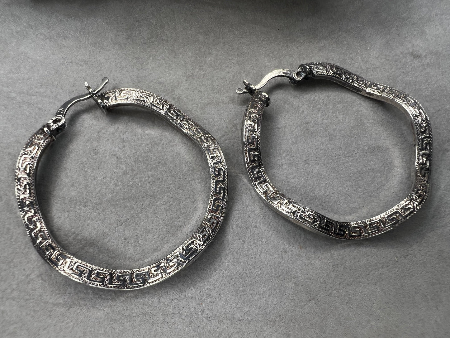 These sterling silver hoop earrings showcase a wavy design enhanced by a timeless Greek key pattern. They combine classic motifs with modern elegance, making them a versatile accessory.