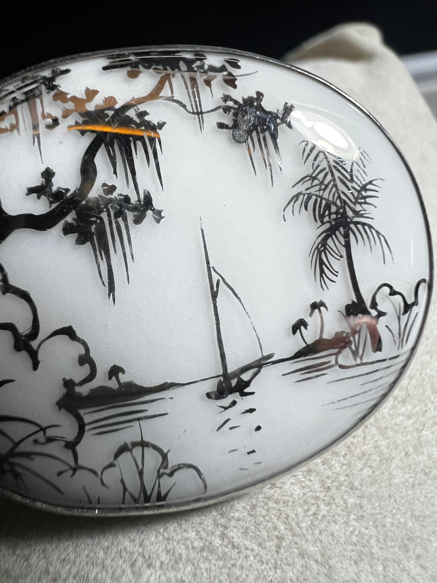 This vintage brooch is an original “cameona” by Florida artist Olive Commons, featuring a hand-painted porcelain scene of a sailboat and palms in her signature black linework with metallic accents. Commons coined the term cameona for these miniature painted landscapes, which became cherished Florida souvenirs and remain highly collectible today.