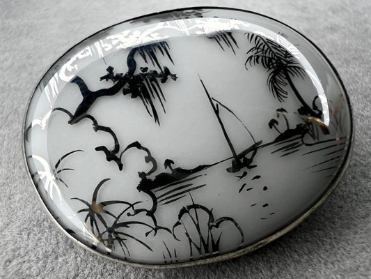 This vintage brooch is an original “cameona” by Florida artist Olive Commons, featuring a hand-painted porcelain scene of a sailboat and palms in her signature black linework with metallic accents. Commons coined the term cameona for these miniature painted landscapes, which became cherished Florida souvenirs and remain highly collectible today.