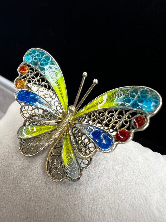 This vintage .800 silver butterfly brooch features a delicate filigree design with a wingspan of 1 5/8" and vibrant enameled wings in a bright mix of colors. Finished with a subtle gold wash, it adds a cheerful touch to any outfit.