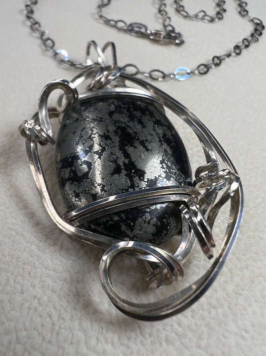 This striking pendant on a chain necklace features a polished Larvikite cabochon, showcasing its distinctive dark gray body with shimmering silver flashes. The stone is set in an artistic sterling silver wirework design, creating a bold and modern statement piece.
