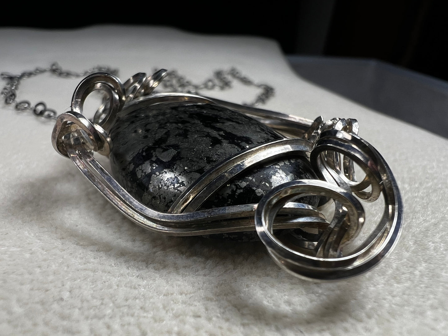 This striking pendant on a chain necklace features a polished Larvikite cabochon, showcasing its distinctive dark gray body with shimmering silver flashes. The stone is set in an artistic sterling silver wirework design, creating a bold and modern statement piece.