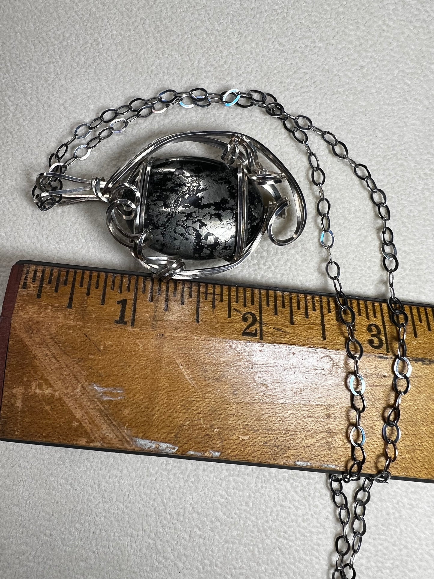 This striking pendant on a chain necklace features a polished Larvikite cabochon, showcasing its distinctive dark gray body with shimmering silver flashes. The stone is set in an artistic sterling silver wirework design, creating a bold and modern statement piece.