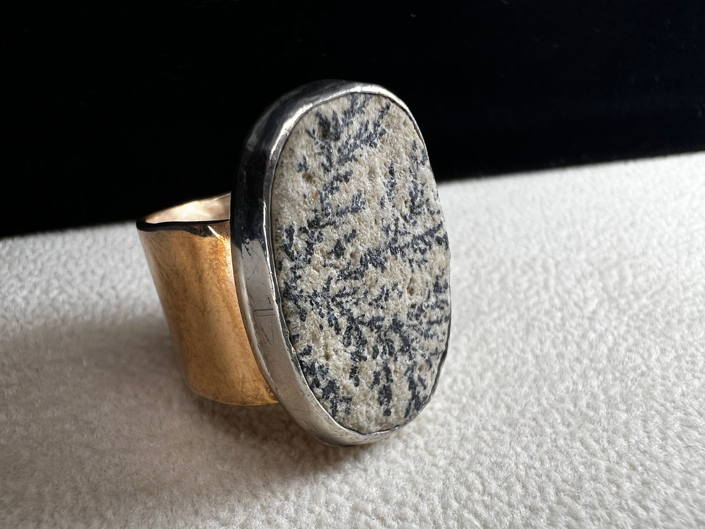 This ring by Loving Anvil features a dendritic limestone cabochon, which displays striking fern-like black inclusions that resemble a natural landscape. The stone is set in sterling silver against a brass band with a thin overlay of gold.