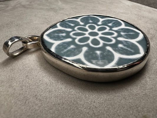 This pendant features a 1.5-inch ceramic disk decorated with a blue and white floral design, framed in sterling silver. Its solid construction makes it a striking piece to wear on a sturdy chain.