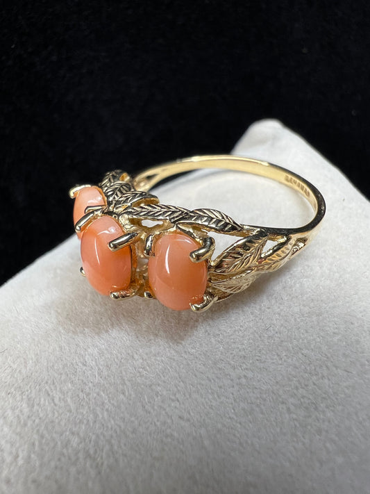This exquisite ring, crafted in the Bahamas from 14 karat gold, features three peachy-salmon coral cabochons elegantly set amid intricately designed gold leaves. The combination of warm coral hues and detailed goldwork creates a distinctive piece.