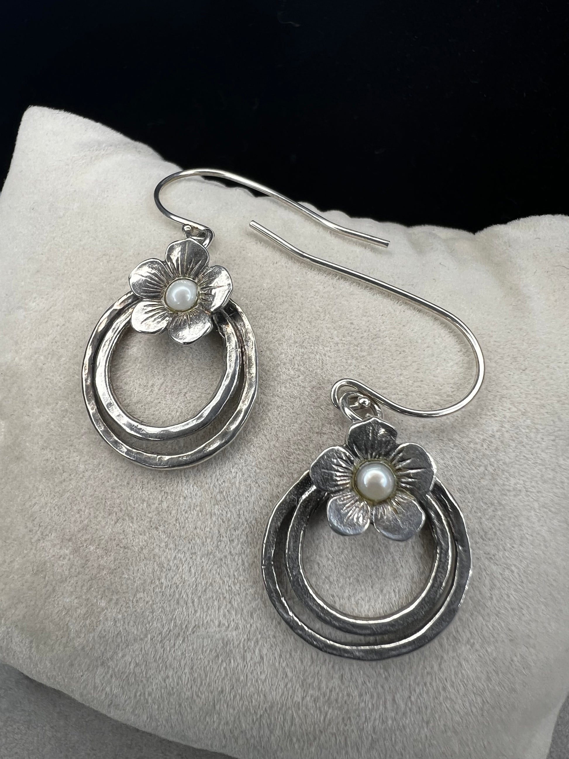 These sterling silver earrings showcase a flower design with a lustrous pearl at its center, elegantly positioned atop a double hoop. The combination offers a refined aesthetic, perfect for adding a sophisticated touch to any ensemble.