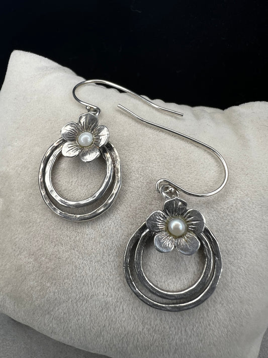 These sterling silver earrings showcase a flower design with a lustrous pearl at its center, elegantly positioned atop a double hoop. The combination offers a refined aesthetic, perfect for adding a sophisticated touch to any ensemble.