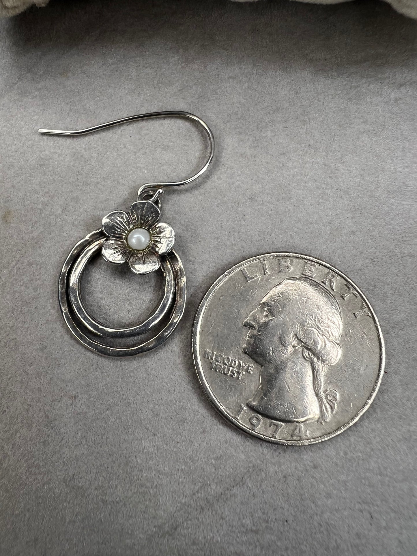 These sterling silver earrings showcase a flower design with a lustrous pearl at its center, elegantly positioned atop a double hoop. The combination offers a refined aesthetic, perfect for adding a sophisticated touch to any ensemble.