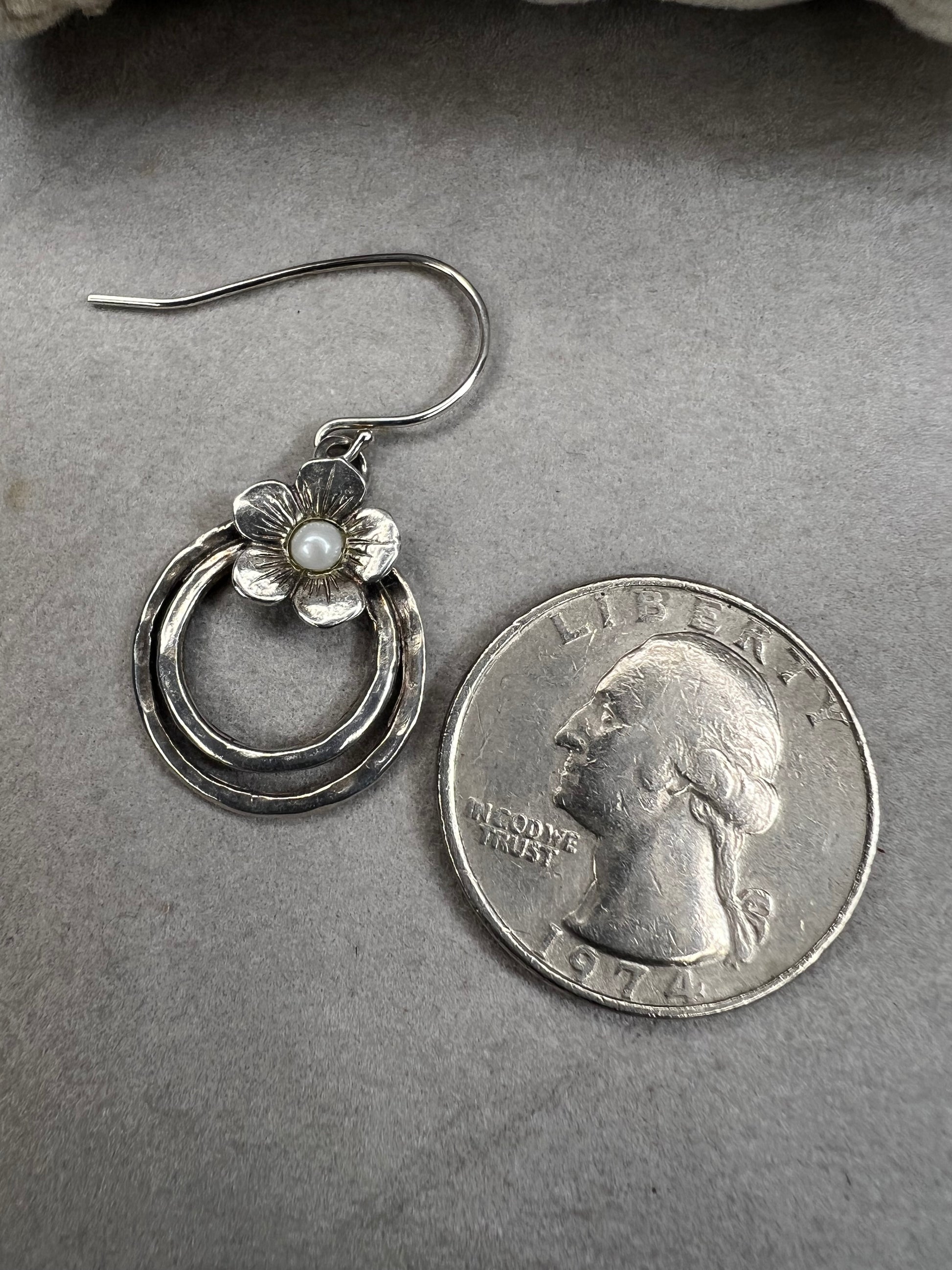 These sterling silver earrings showcase a flower design with a lustrous pearl at its center, elegantly positioned atop a double hoop. The combination offers a refined aesthetic, perfect for adding a sophisticated touch to any ensemble.