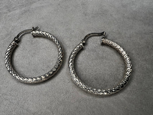 These sterling silver hoop earrings feature a diamond-cut texture that adds subtle shimmer and dynamic movement. Their moderate size balances statement presence with comfortable wearability.