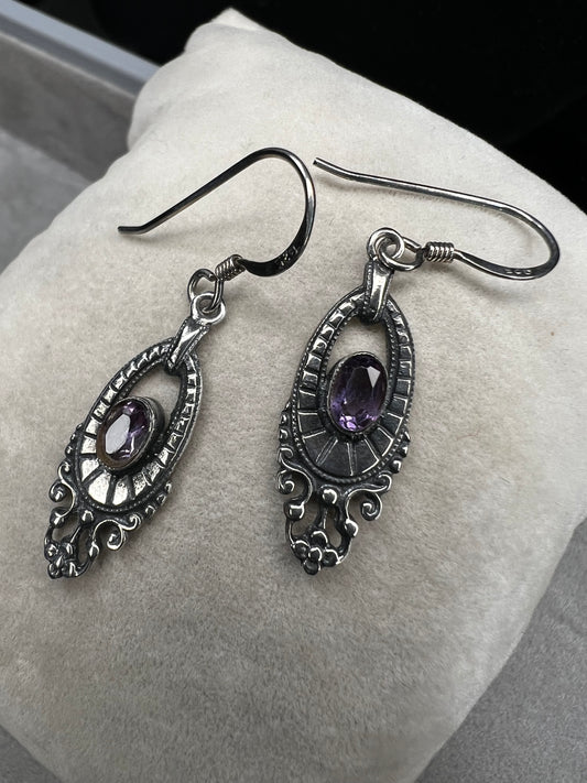 These sterling silver dangle earrings showcase an intricate design centered around a vibrant amethyst gemstone. The rich purple hue of the amethyst will add a refined splash of color to your outfit.