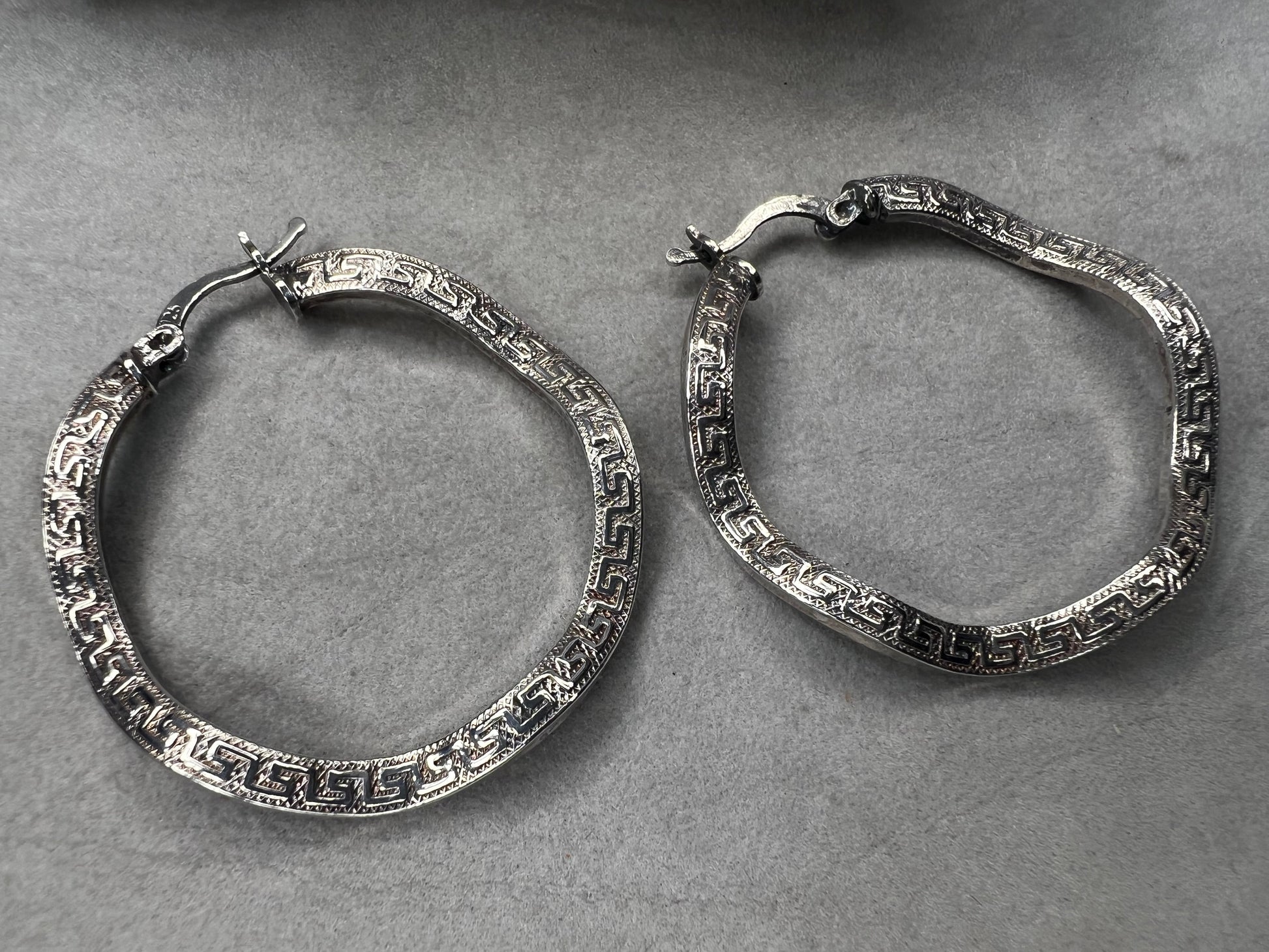 These sterling silver hoop earrings showcase a wavy design enhanced by a timeless Greek key pattern. They combine classic motifs with modern elegance, making them a versatile accessory.