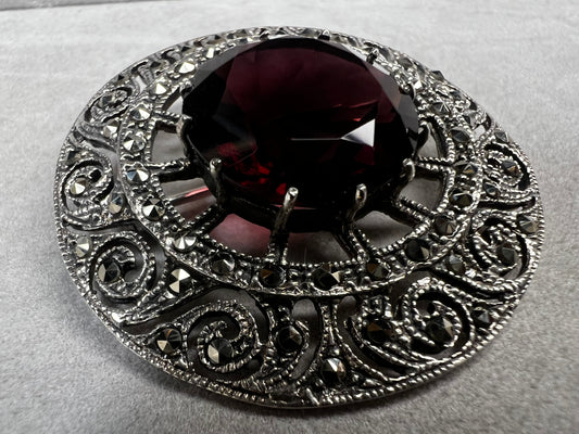 This vintage sterling silver brooch measures 1.5" in diameter and features an elegant open work design. The marcasites catch the light beautifully, while the deep purple faux stone adds a rich, eye-catching touch. Perfect for adding timeless charm to any outfit.