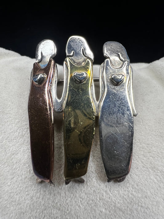 Signed "Far Fetched," this sterling silver pin features three women side by side, each wearing a different colored dress accented in brass, copper, and silver. A heartfelt design, it symbolizes friendship, sisterhood, and unity.