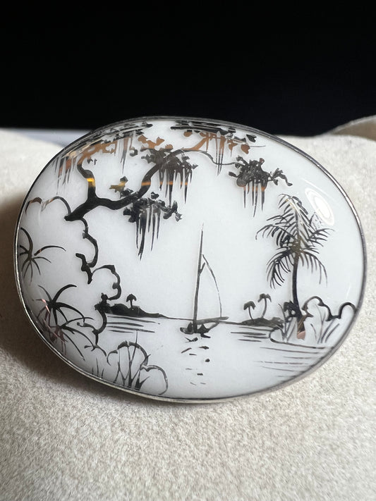 This vintage brooch is an original “cameona” by Florida artist Olive Commons, featuring a hand-painted porcelain scene of a sailboat and palms in her signature black linework with metallic accents. Commons coined the term cameona for these miniature painted landscapes, which became cherished Florida souvenirs and remain highly collectible today.