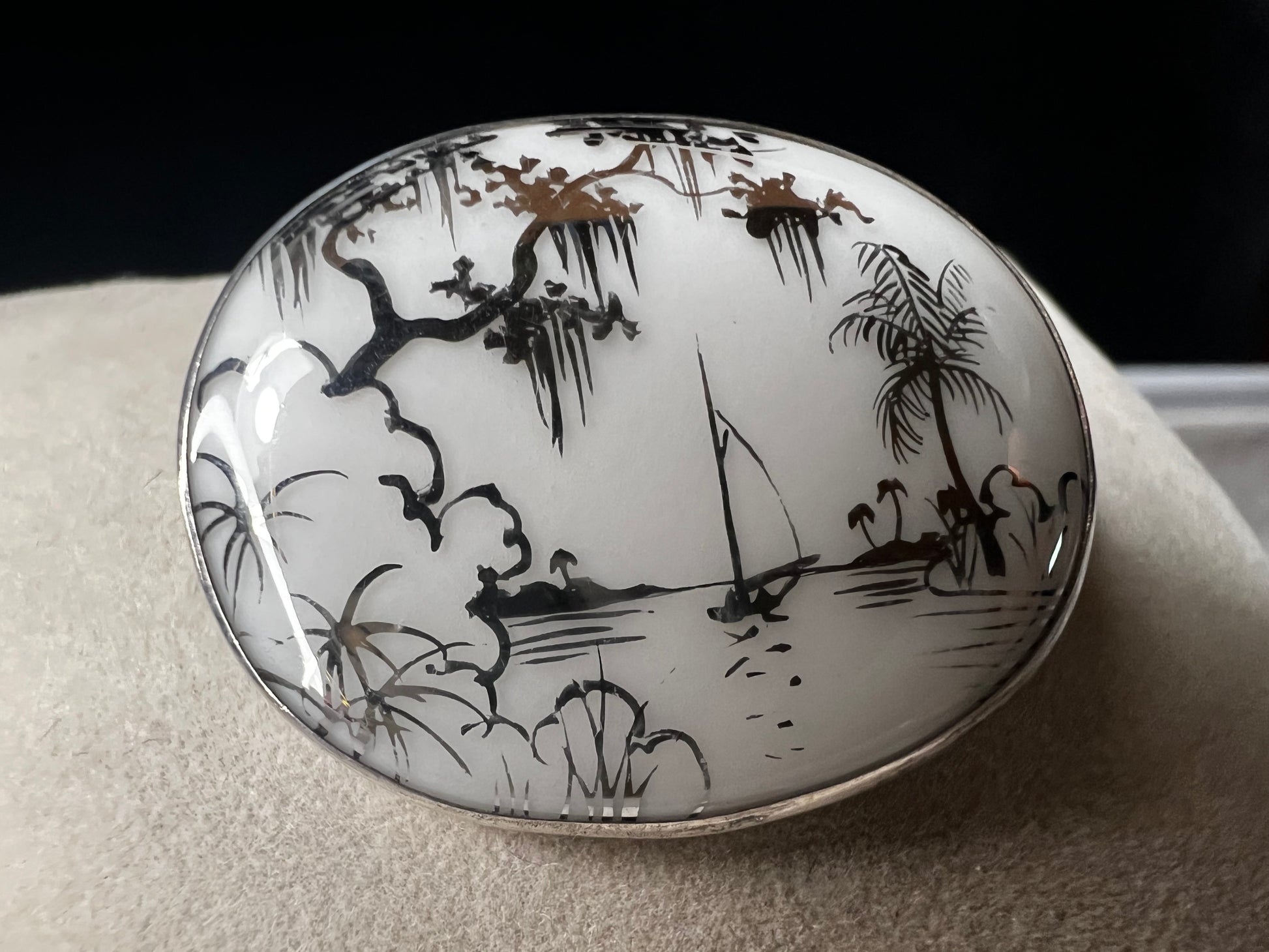 This vintage brooch is an original “cameona” by Florida artist Olive Commons, featuring a hand-painted porcelain scene of a sailboat and palms in her signature black linework with metallic accents. Commons coined the term cameona for these miniature painted landscapes, which became cherished Florida souvenirs and remain highly collectible today.