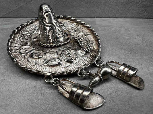 This charming 1950s sterling silver brooch captures a playful scene with a large sombrero, a pair of sandals, and a maraca. Its unique design reflects vintage character and cultural flair, making it a delightful accessory that adds personality and nostalgia to your collection.