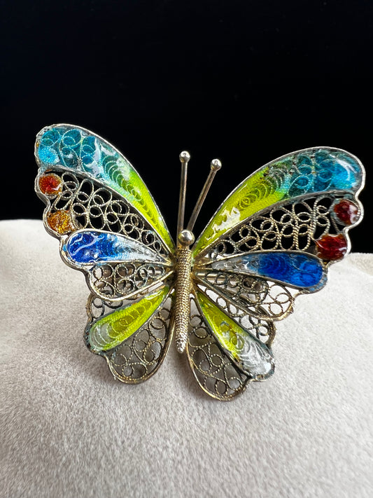 This vintage .800 silver butterfly brooch features a delicate filigree design with a wingspan of 1 5/8" and vibrant enameled wings in a bright mix of colors. Finished with a subtle gold wash, it adds a cheerful touch to any outfit.