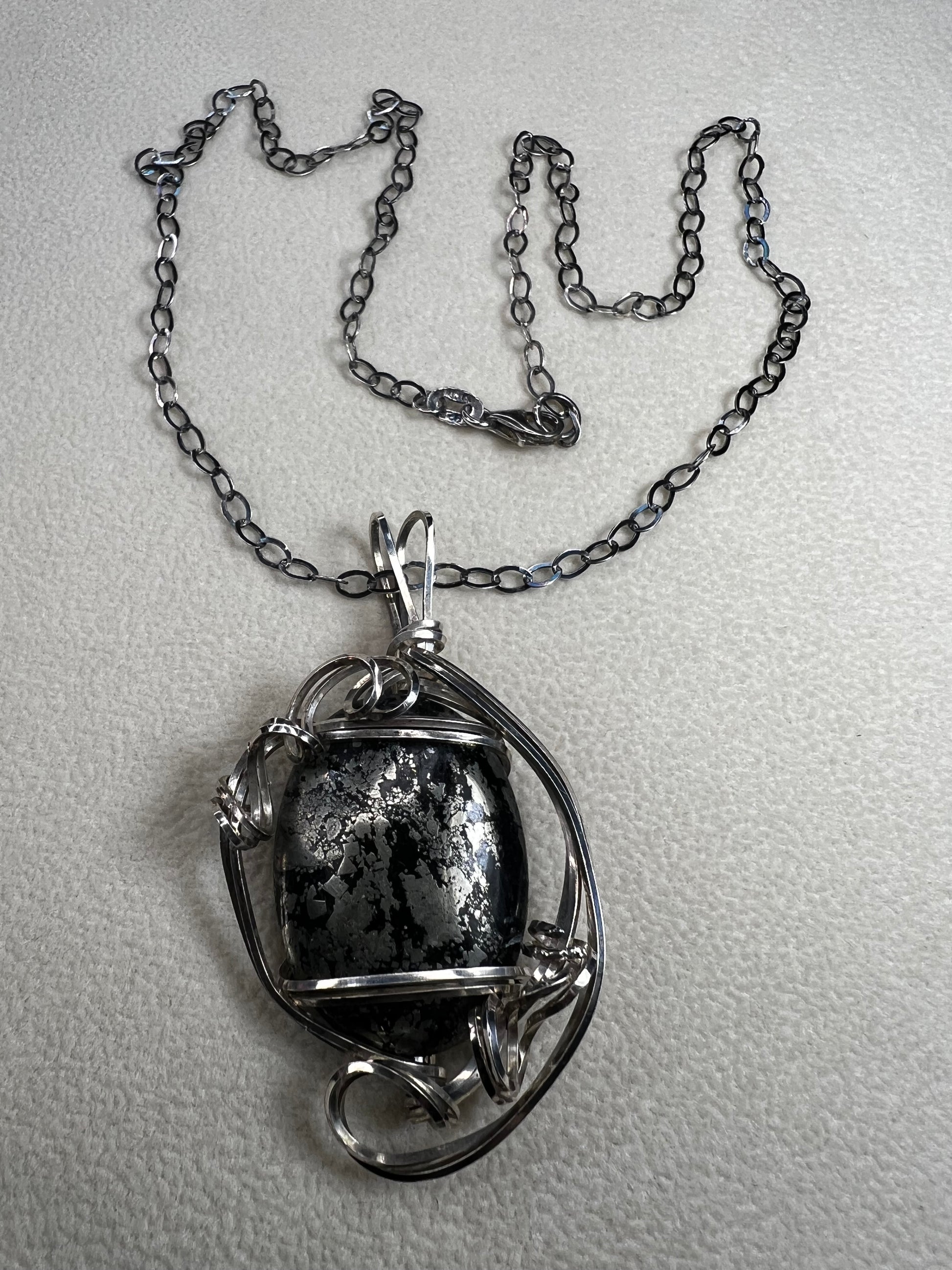 This striking pendant on a chain necklace features a polished Larvikite cabochon, showcasing its distinctive dark gray body with shimmering silver flashes. The stone is set in an artistic sterling silver wirework design, creating a bold and modern statement piece.