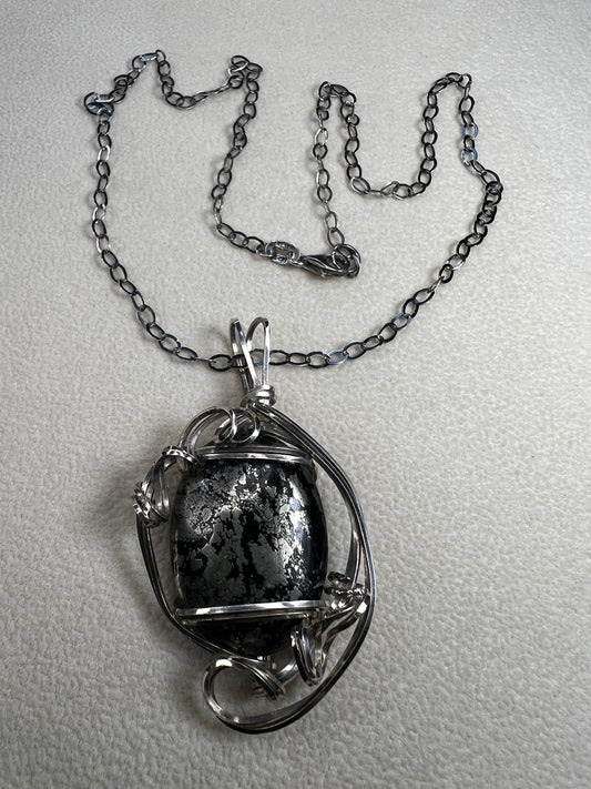 This striking pendant on a chain necklace features a polished Larvikite cabochon, showcasing its distinctive dark gray body with shimmering silver flashes. The stone is set in an artistic sterling silver wirework design, creating a bold and modern statement piece.