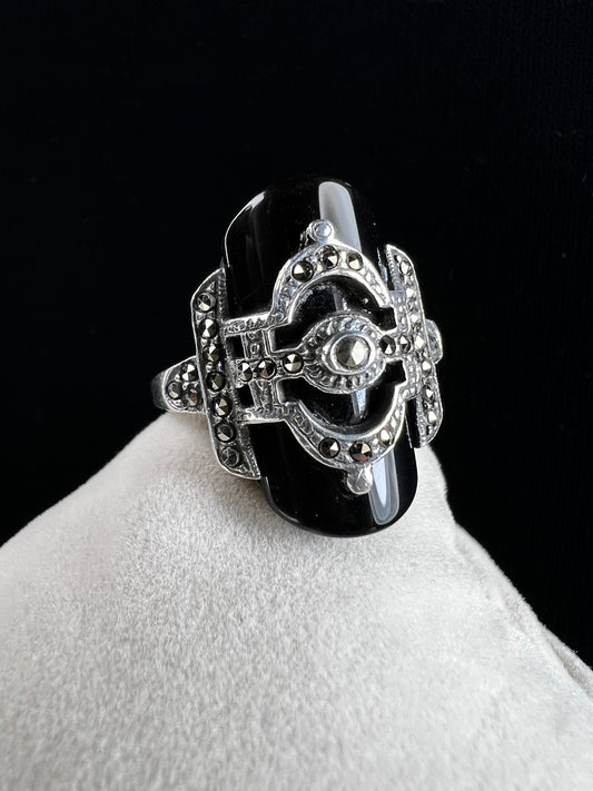 This striking vintage sterling silver ring features a polished black onyx cabochon accented with sparkling marcasite. Designed in an Art Deco revival style, this bold piece blends timeless elegance with dramatic flair.