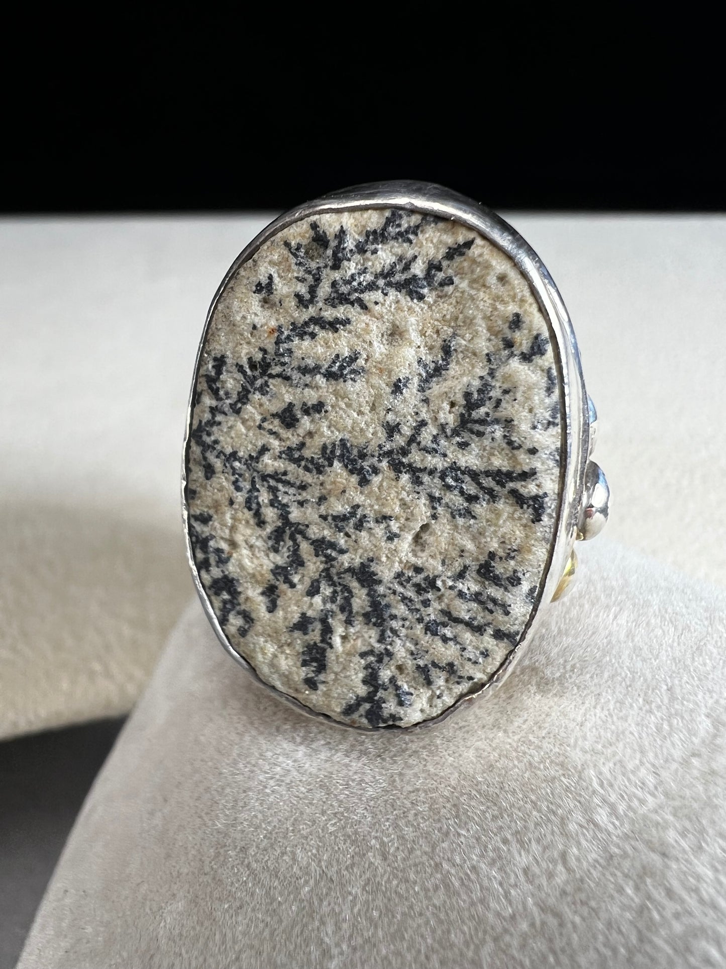 This ring by Loving Anvil features a dendritic limestone cabochon, which displays striking fern-like black inclusions that resemble a natural landscape. The stone is set in sterling silver against a brass band with a thin overlay of gold.