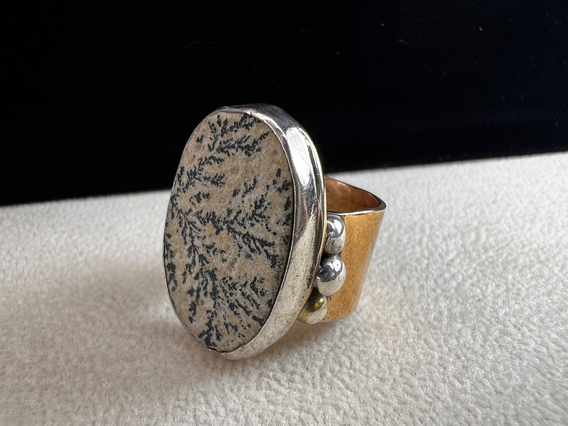 This ring by Loving Anvil features a dendritic limestone cabochon, which displays striking fern-like black inclusions that resemble a natural landscape. The stone is set in sterling silver against a brass band with a thin overlay of gold.