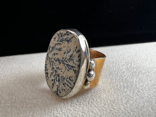 This ring by Loving Anvil features a dendritic limestone cabochon, which displays striking fern-like black inclusions that resemble a natural landscape. The stone is set in sterling silver against a brass band with a thin overlay of gold.