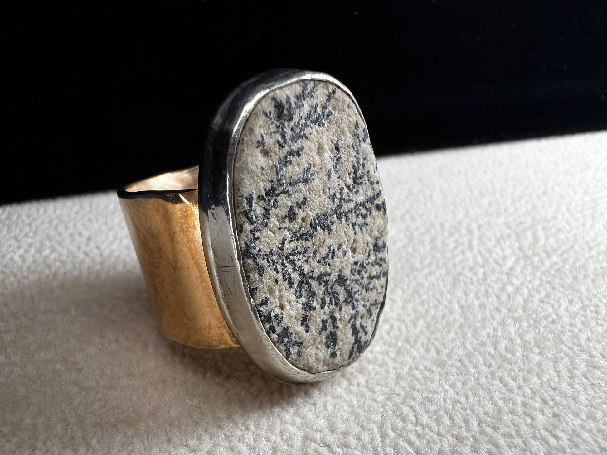 This ring by Loving Anvil features a dendritic limestone cabochon, which displays striking fern-like black inclusions that resemble a natural landscape. The stone is set in sterling silver against a brass band with a thin overlay of gold.
