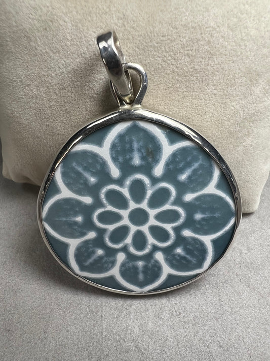 This pendant features a 1.5-inch ceramic disk decorated with a blue and white floral design, framed in sterling silver. Its solid construction makes it a striking piece to wear on a sturdy chain.
