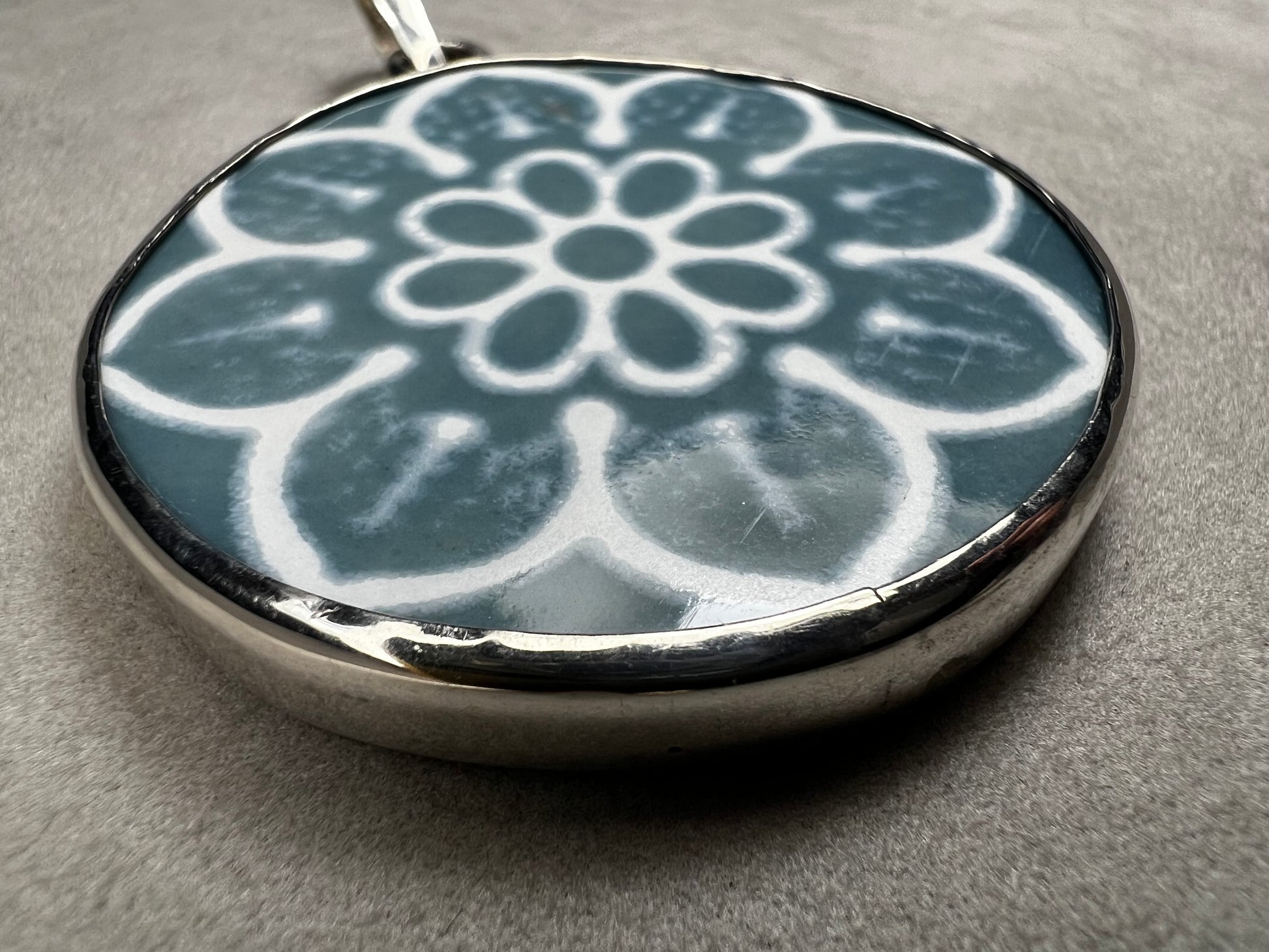 This pendant features a 1.5-inch ceramic disk decorated with a blue and white floral design, framed in sterling silver. Its solid construction makes it a striking piece to wear on a sturdy chain.