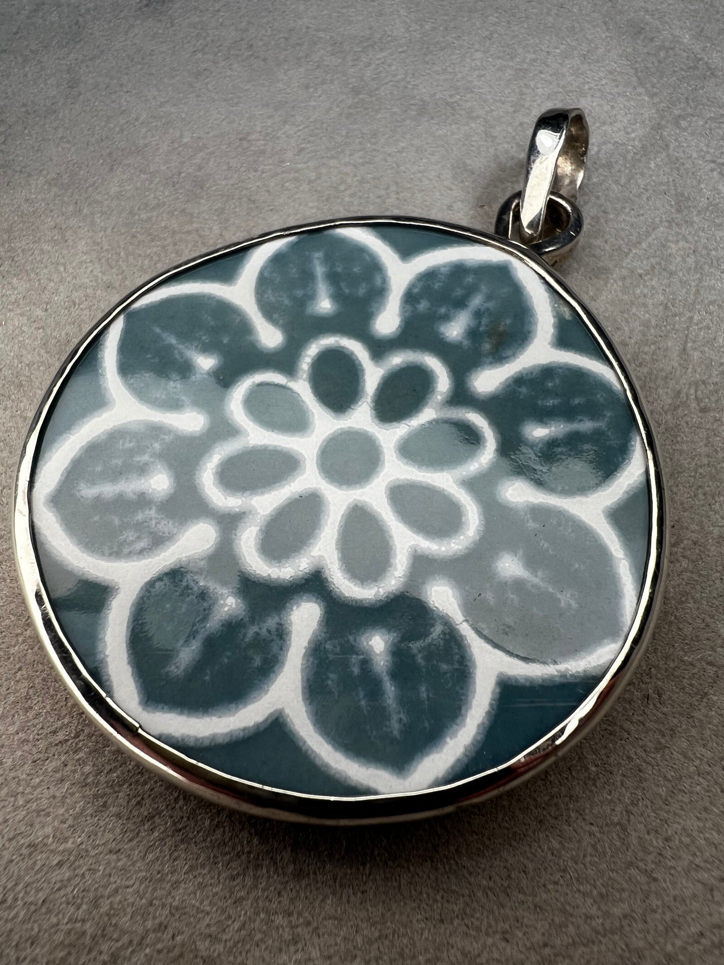 This pendant features a 1.5-inch ceramic disk decorated with a blue and white floral design, framed in sterling silver. Its solid construction makes it a striking piece to wear on a sturdy chain.