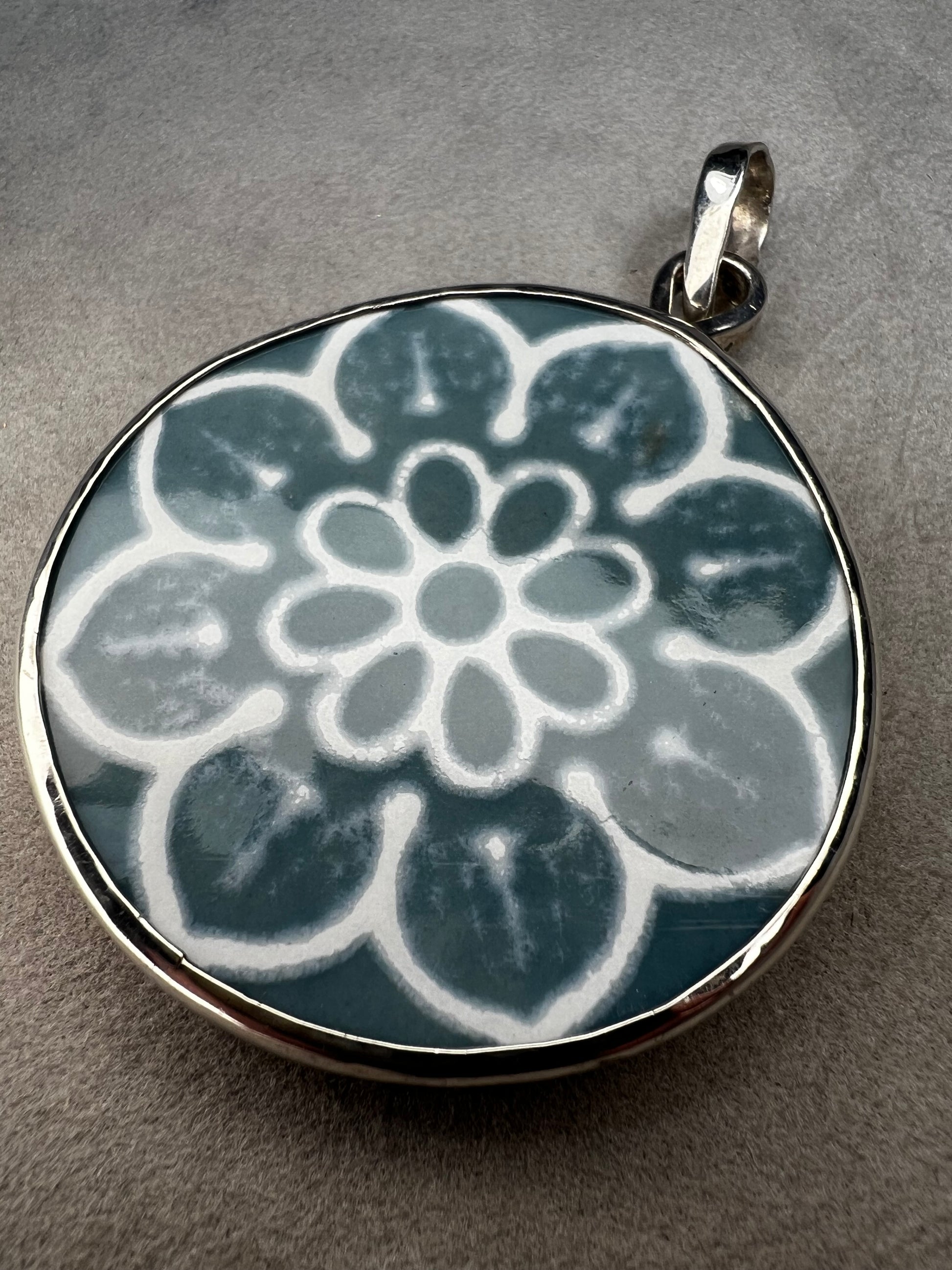 This pendant features a 1.5-inch ceramic disk decorated with a blue and white floral design, framed in sterling silver. Its solid construction makes it a striking piece to wear on a sturdy chain.
