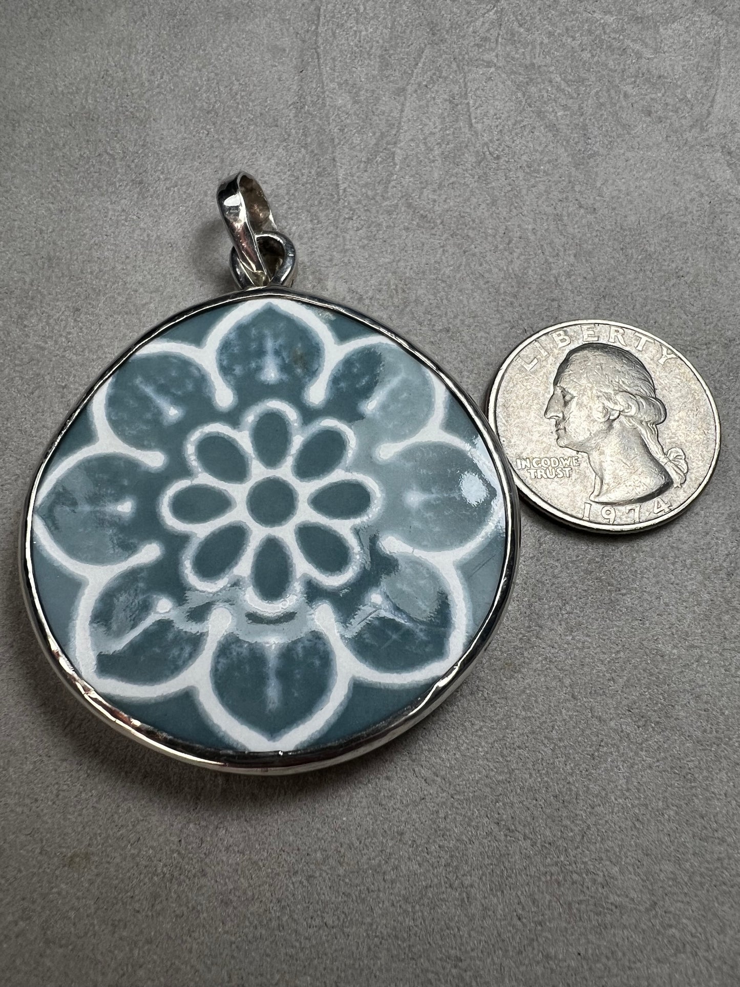 This pendant features a 1.5-inch ceramic disk decorated with a blue and white floral design, framed in sterling silver. Its solid construction makes it a striking piece to wear on a sturdy chain.