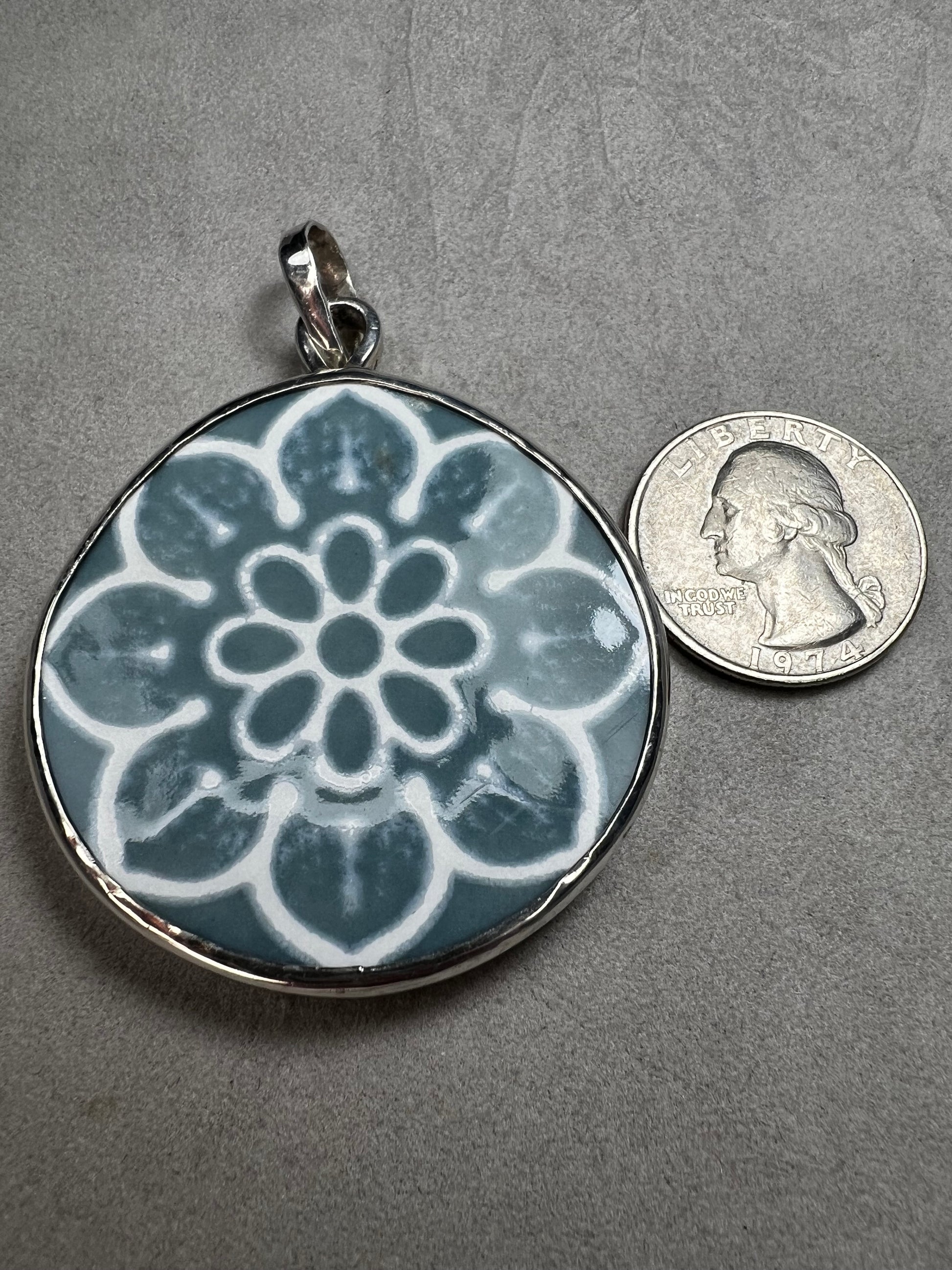 This pendant features a 1.5-inch ceramic disk decorated with a blue and white floral design, framed in sterling silver. Its solid construction makes it a striking piece to wear on a sturdy chain.